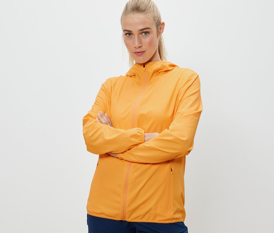 Frau posiert mit verschränkten Armen in orangefarbener, packbarer Funktionsjacke.