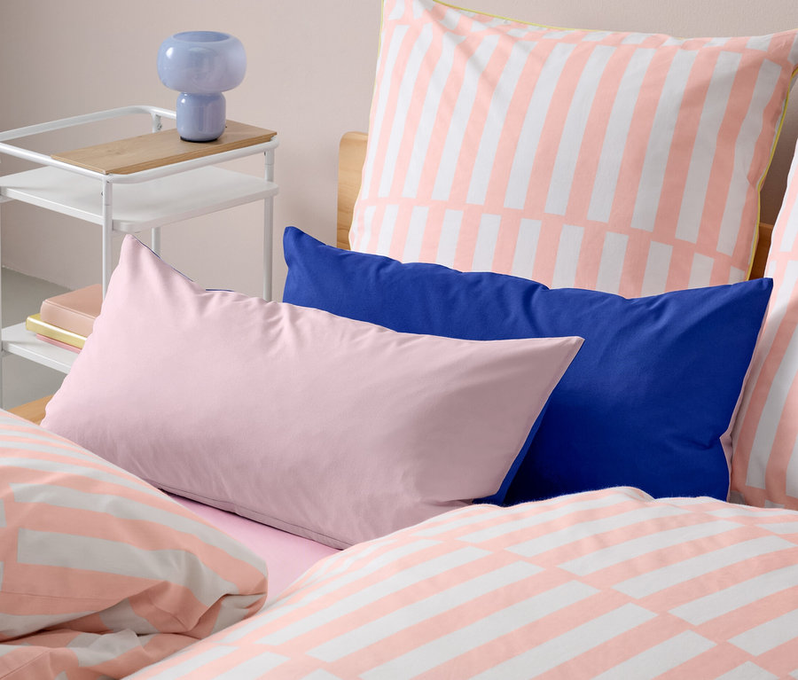 Zwei dunkelblau und rosa Renforcé-Kopfkissenbezüge liegen auf einem Bett mit gestreifter Bettwäsche. Neben dem Bett steht ein Tisch mit einer Lampe.