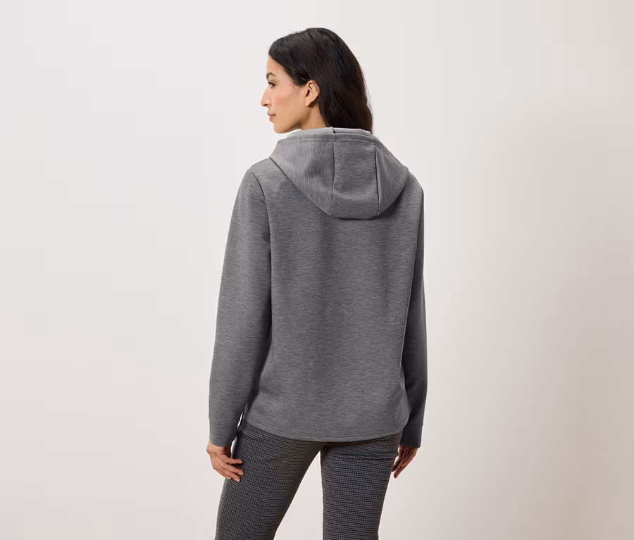 Rückansicht einer Frau mit grauem Kapuzenpullover und gemusterter Hose.