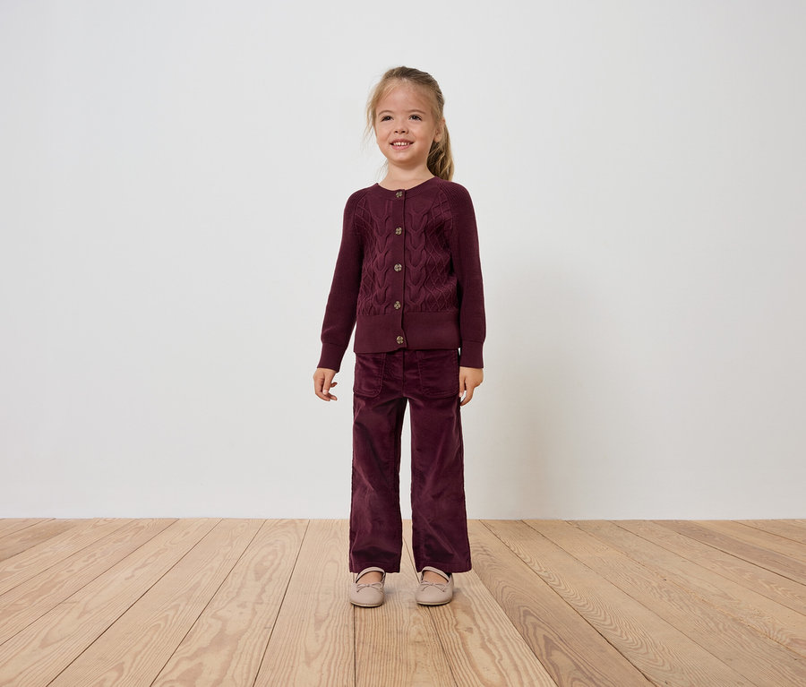Ein lächelndes Mädchen in einem burgunderroten Samt-Cardigan und einer Hose steht auf einem Holzboden.