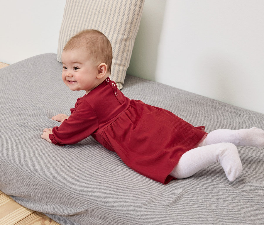 Baby in rotem Kleid und weißen Strumpfhosen liegt auf einer grauen Unterlage.