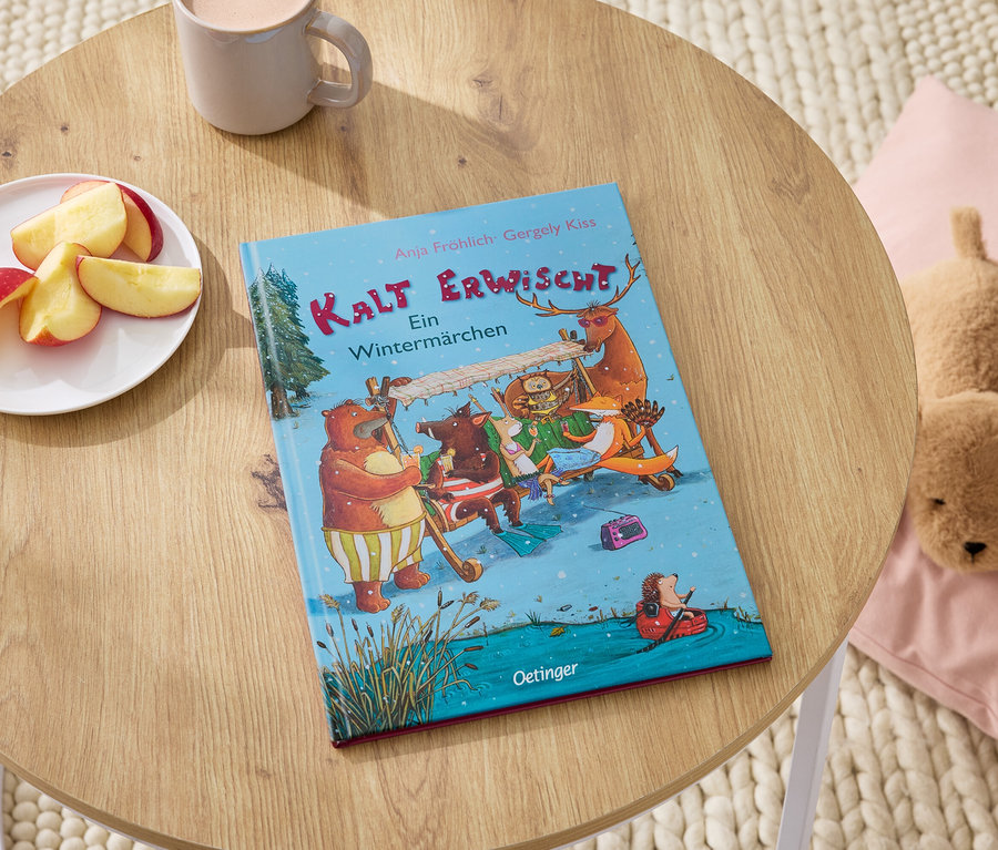 Auf einem Holztisch liegen das Buch "Kalt Erwischt", ein Teller mit Apfelspalten, eine Tasse und ein Plüschhund auf einem rosa Kissen.