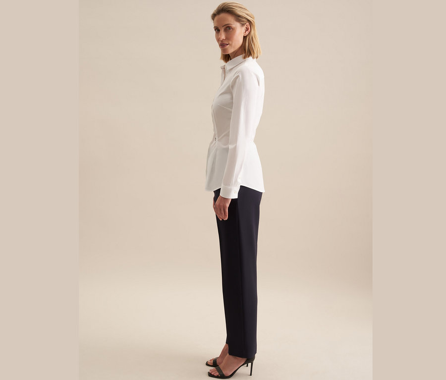 Frau steht seitlich und trägt eine weisse Seidensticker Slimfit Bluse und dunkelblaue Hosen.