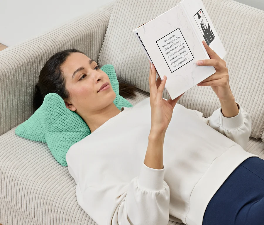 Eine Frau liegt auf einem Sofa und liest ein Buch, ihr Kopf ruht auf einem Lesekissen.