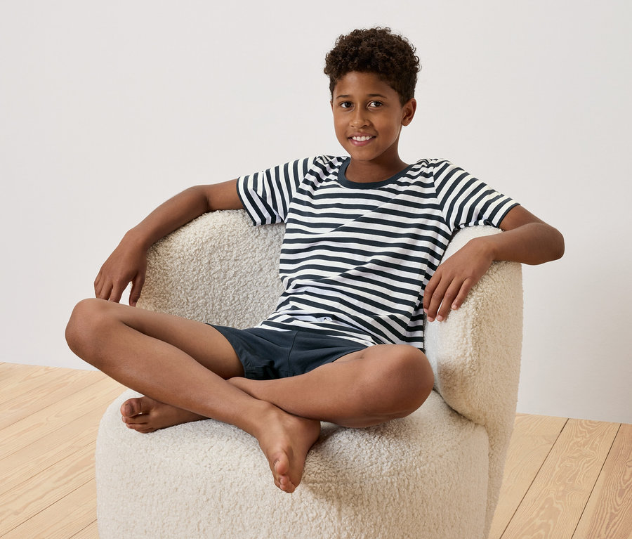 Ein junger Mann sitzt mit gekreuzten Beinen in einem cremefarbenen Sessel. Er trägt ein gestreiftes T-Shirt und marineblaue Shorts.