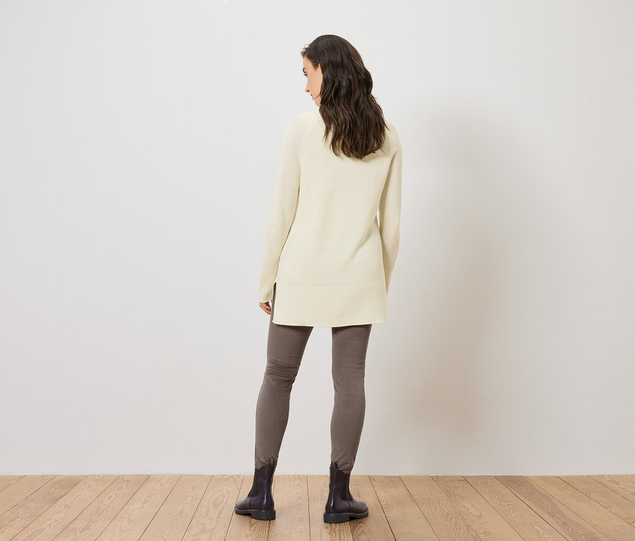 Frau von hinten trägt einen langen Strickpullover in Creme, graue Leggings und schwarze Stiefel.