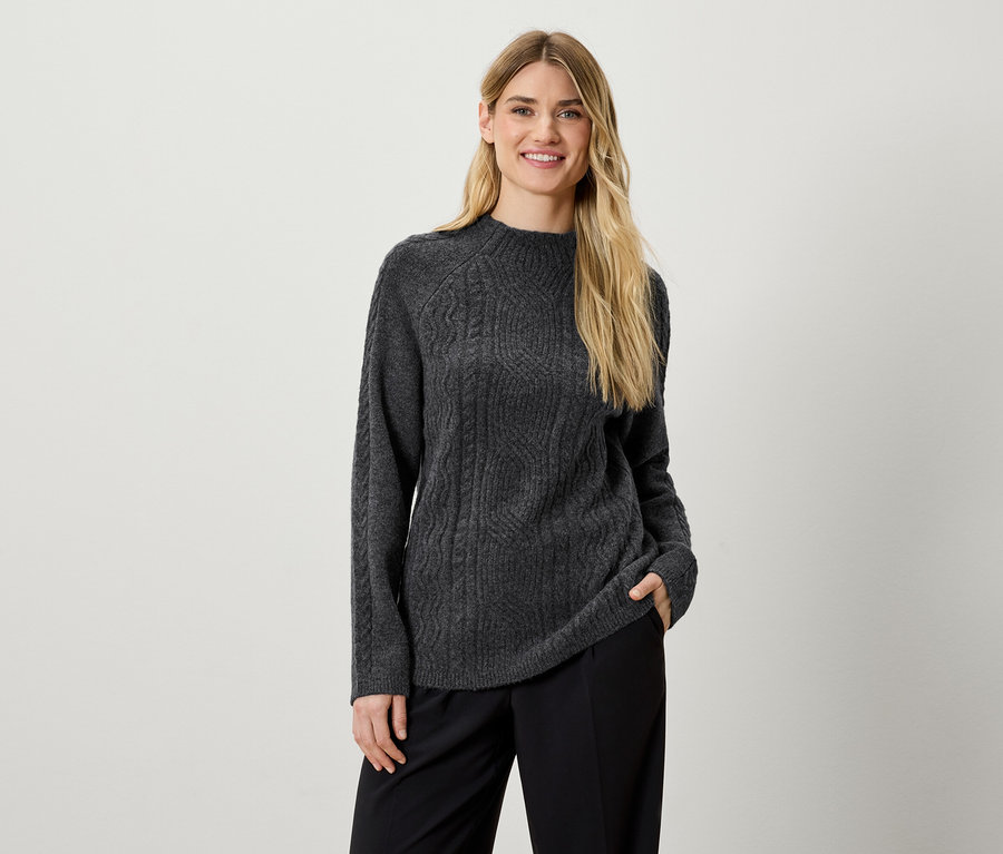 Eine Frau posiert in einem grauen Strickpullover und einer schwarzen Hose.