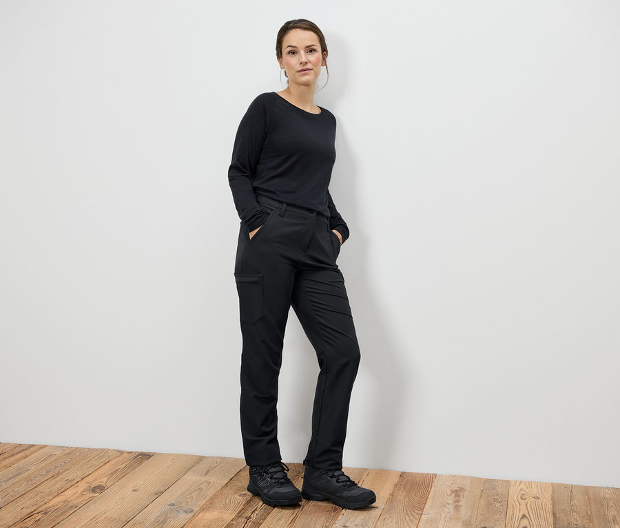 Frau steht mit Händen in den Taschen in schwarzen Hosen und schwarzen Stiefeln.