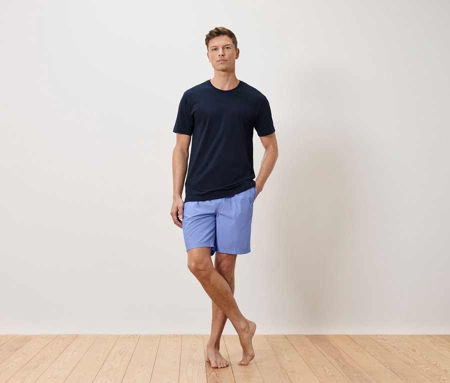 Mann mit marineblauem T-Shirt und hellblauen Shorts, stehend vor einer weißen Wand.
