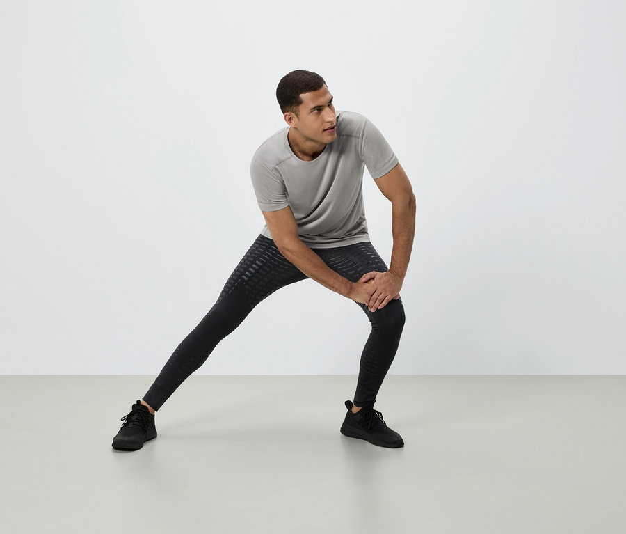 Ein Mann dehnt sich in einem grauen T-Shirt, schwarzen Leggings und schwarzen Turnschuhen vor einem weißen Hintergrund.