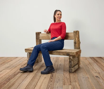 Frau sitzt auf einer Holzbank, trägt eine rote Bluse, blaue Hosen und Stiefel.