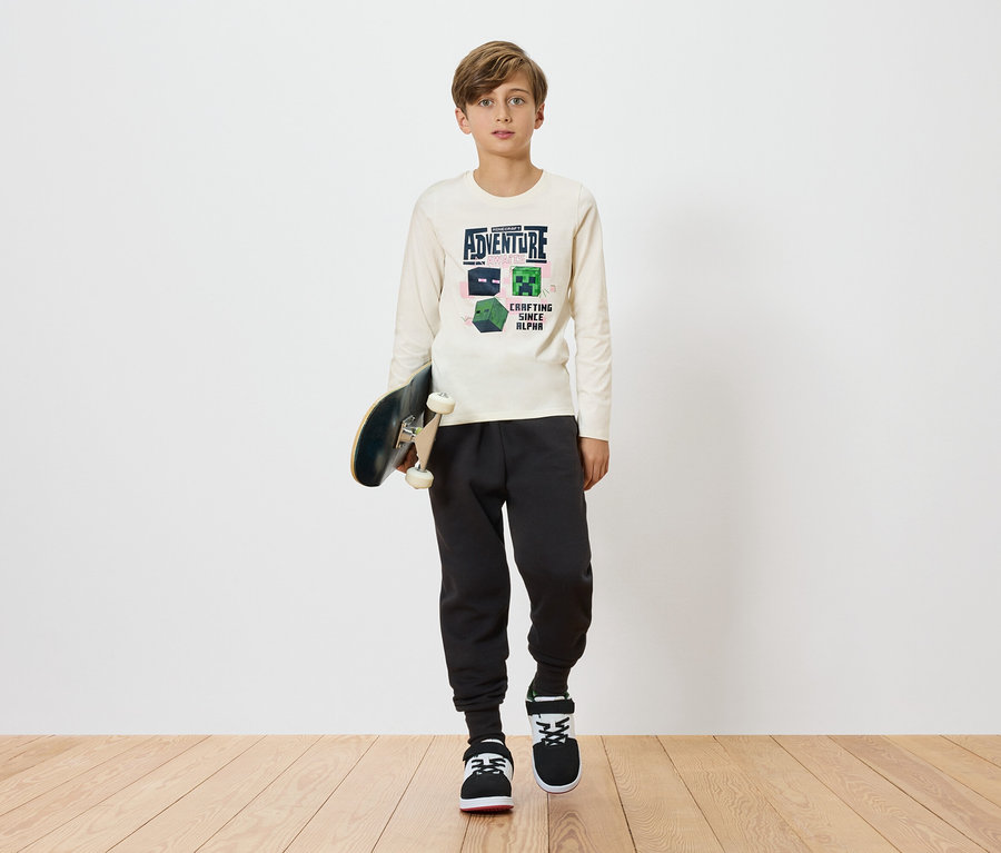 Ein Junge hält ein Skateboard und trägt ein helles Langarmshirt mit einem Minecraft-Design, kombiniert mit dunklen Jogginghosen und Turnschuhen vor einem schlichten Hintergrund.