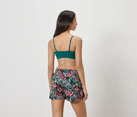 Eine Frau steht mit dem Rücken zur Kamera, sie trägt einen grünen BH und kurze, blumige Shorts.