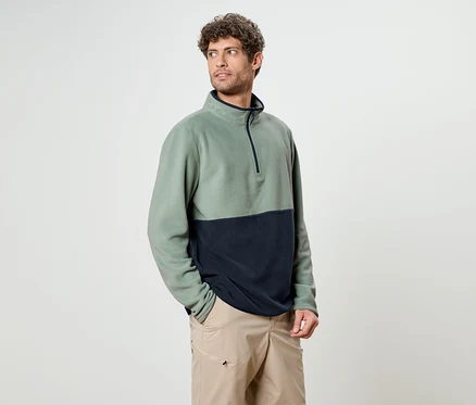 Ein Mann mit lockigem Haar trägt ein zweifarbiges Fleece-Oberteil in Grün und Marineblau mit beigen Shorts.