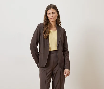 Frau in braunem Blazer und Hose mit gelbem Oberteil vor hellem Hintergrund.