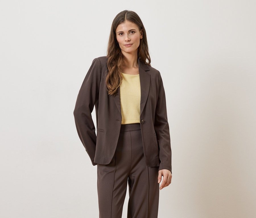 Frau in braunem Blazer und Hose mit gelbem Oberteil vor hellem Hintergrund.