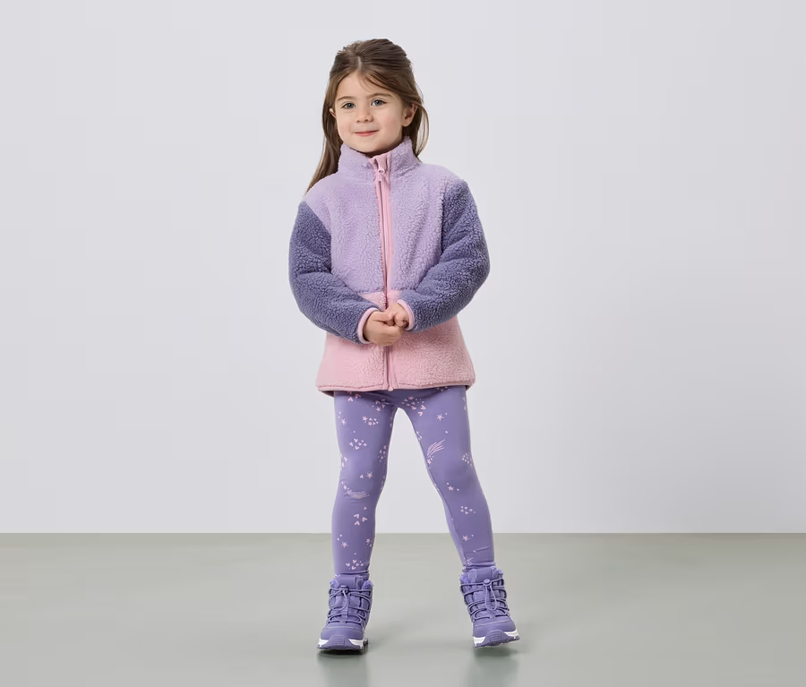Ein Mädchen steht mit verschränkten Händen da und trägt eine Fleecejacke in Lila und Rosa, Leggings mit Sternen und lila Stiefel.