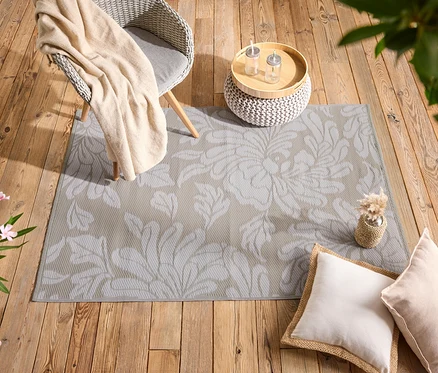 Ein beiger Teppich mit Blumenmuster liegt auf einem Holzboden, daneben Kissen, ein Stuhl und ein Tisch.