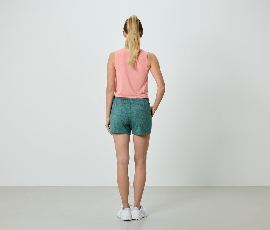 Rückansicht einer Frau mit blonden Haaren in einem lachsfarbenen Tanktop und grünen Shorts, die in weißen Turnschuhen vor einem neutralen Hintergrund steht.