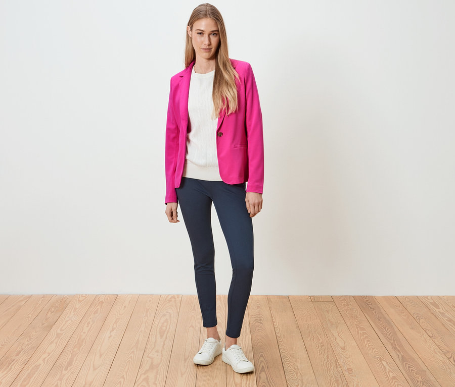 Frau steht und trägt einen pinken Blazer, ein weißes Oberteil, blaue Leggings und weiße Turnschuhe.