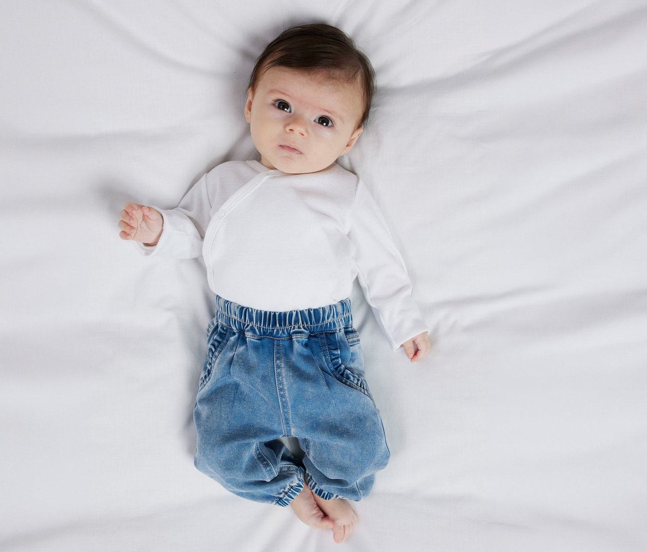 Baby liegt auf dem Rücken und trägt eine weiße Wickelbluse und eine NAME IT Baby-Jeans.