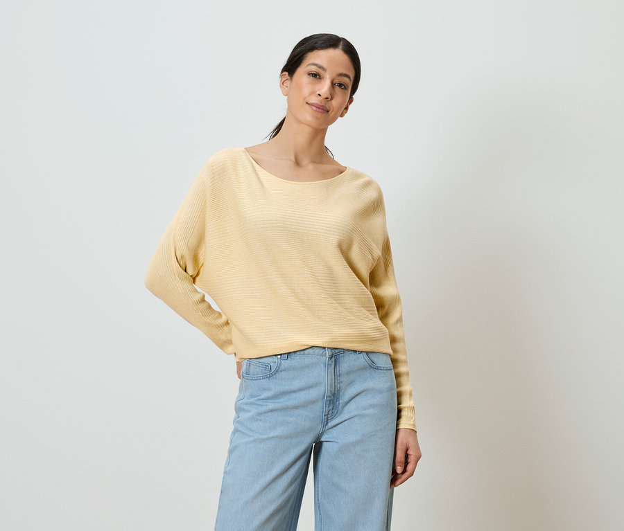 Frau mit gelbem Pullover und blauen Jeans posiert vor einem weißen Hintergrund.