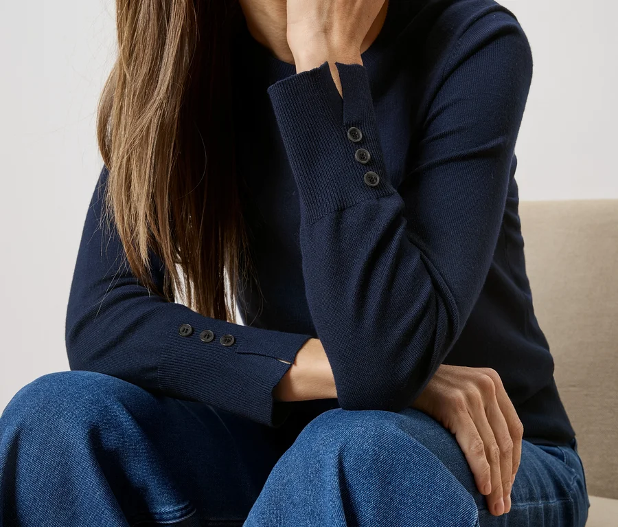 Frau sitzt in marineblauem Pullover mit Knöpfen am Ärmel und blauen Jeans auf einem Stuhl.