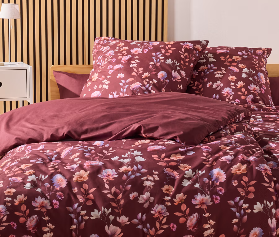 Burgunderfarbene Bettwäsche mit Blumenmuster liegt auf einem Bett mit Holzkopfende vor einer Wand mit Holzpaneelen.