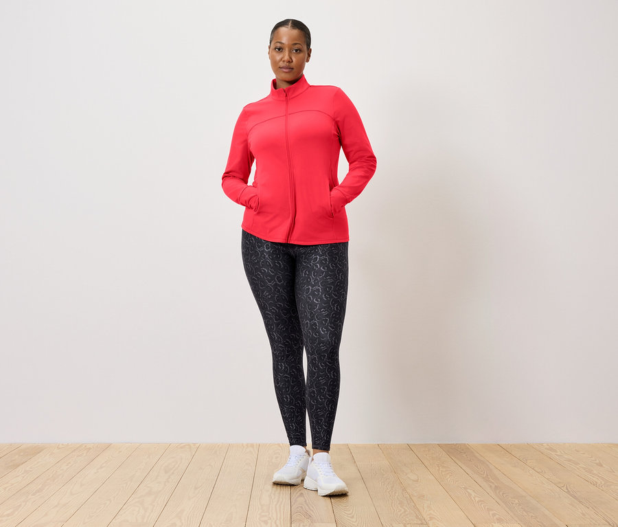 Frau mit roter Jacke und gemusterten Leggings posiert vor einer Wand.