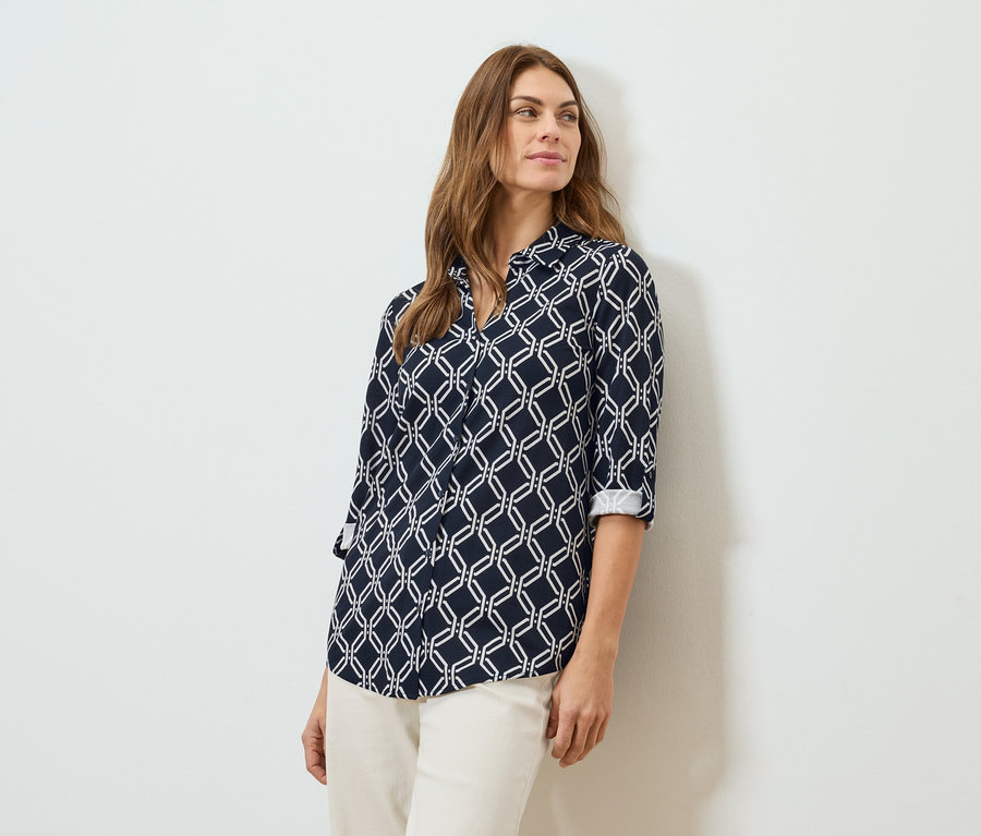 Frau mit braunen Haaren, die eine marineblaue, geometrisch gemusterte Bluse und eine cremefarbene Hose trägt.