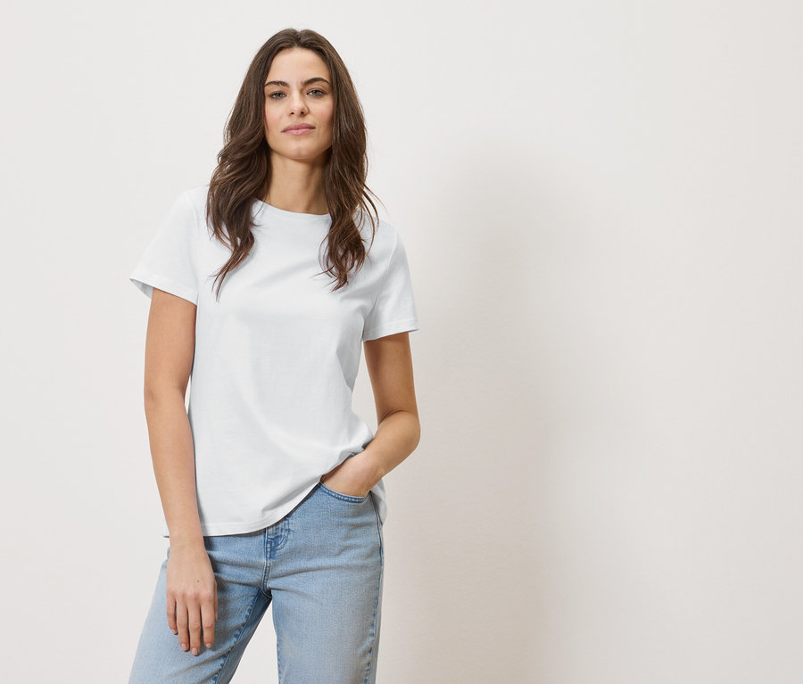 Frau mit braunen Haaren trägt ein weißes T-Shirt und Jeans