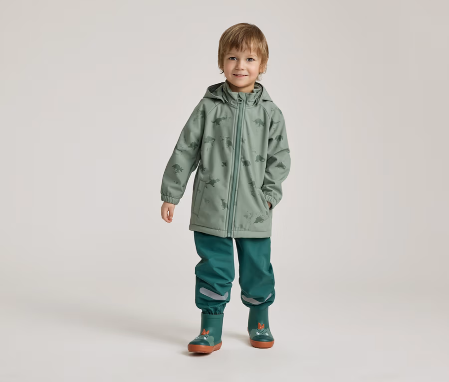 Ein Junge steht in grüner Dino-Regenjacke, grüner Hose und Gummistiefeln vor einem hellen Hintergrund.