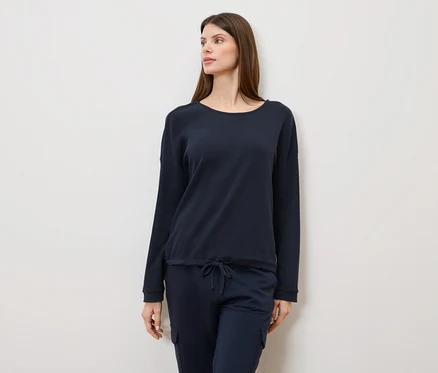 Frau mit langen braunen Haaren, die einen marineblauen Pullover und eine marineblaue Hose trägt und zur Seite schaut.