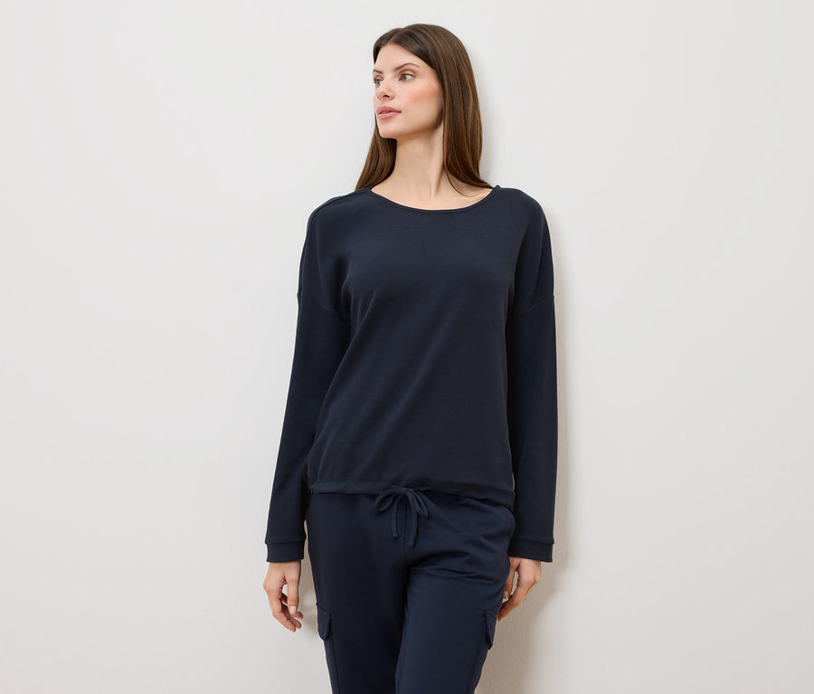 Frau mit langen braunen Haaren, die einen marineblauen Pullover und eine marineblaue Hose trägt und zur Seite schaut.