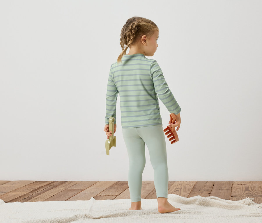 Ein junges Mädchen steht mit dem Rücken zur Kamera, trägt ein gestreiftes Oberteil und Leggings und hält Spielzeug in den Händen.