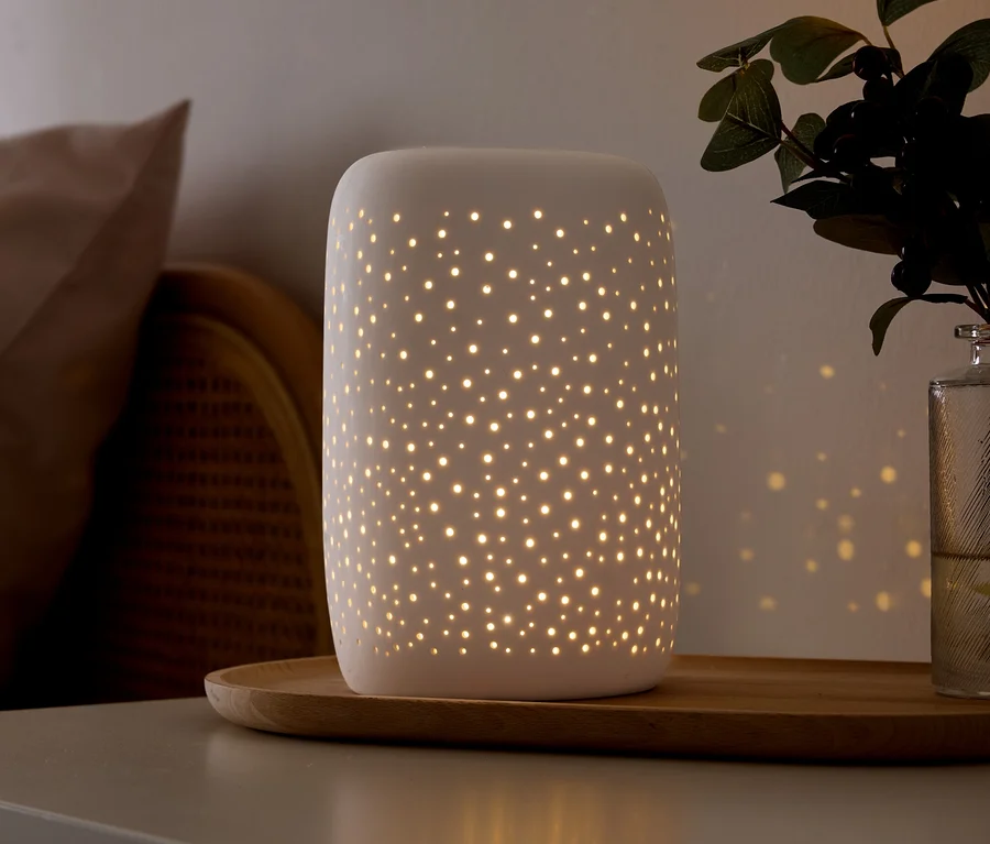Eine weiße Keramiklampe mit kleinen Perforationen strahlt warmes Licht aus und wirft sternförmige Muster an die Wand. Sie steht auf einem Holztablett auf einer grauen Oberfläche, neben einem beigen Kissen, Korbmöbeln und einer grünen Pflanze in einer Glasvase.