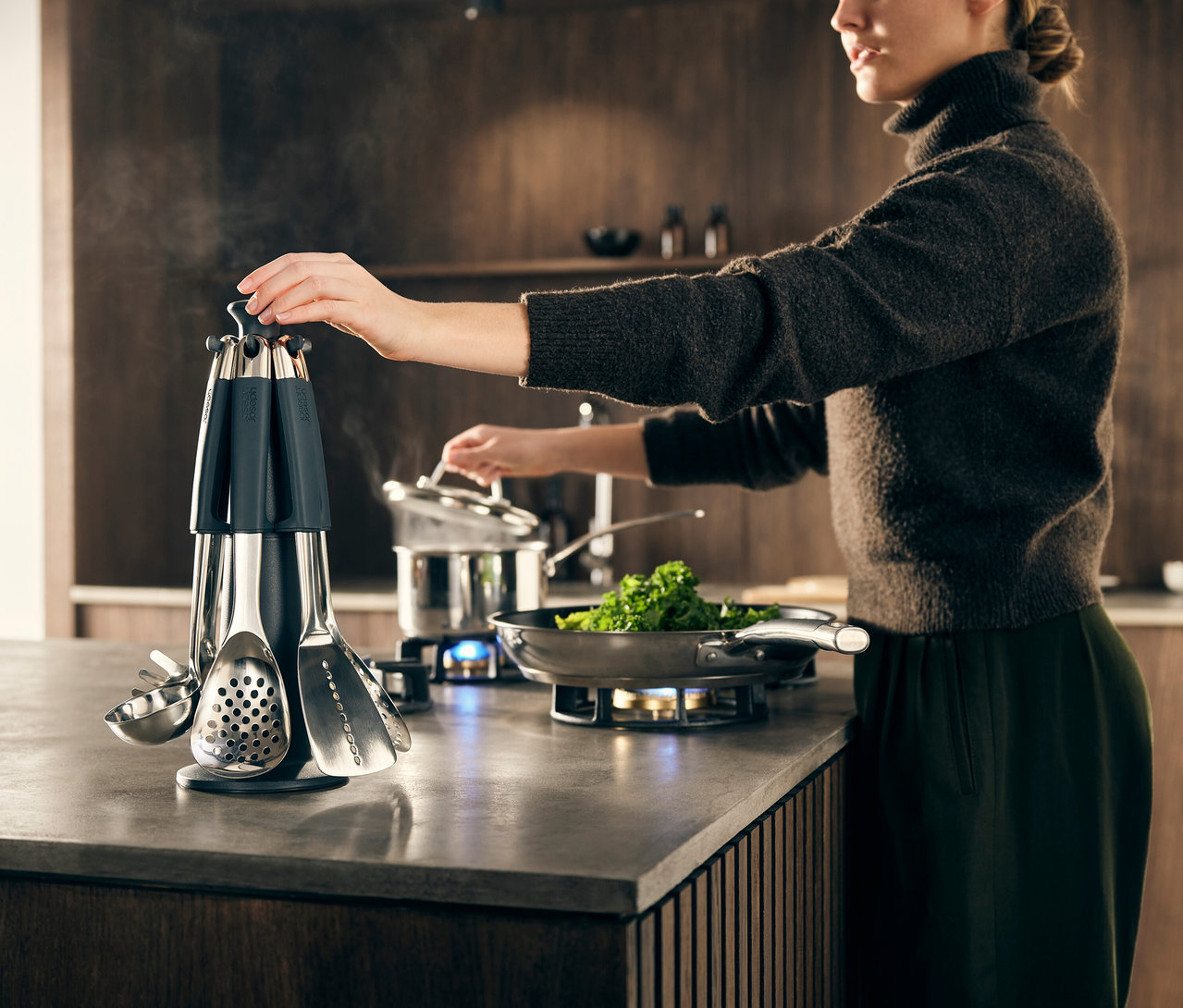 Frau greift nach einem Utensil am Joseph Joseph Elevate Steel Carousel, während sie Gemüse in einer Pfanne und einem Topf auf einem Gasherd kocht.