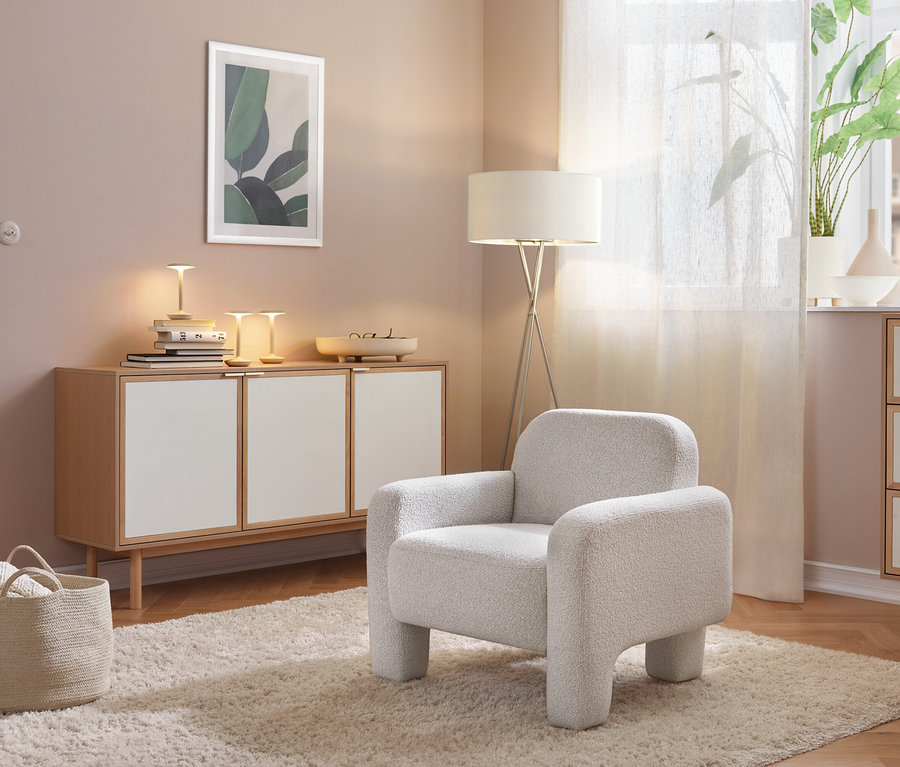 Ein gemütlicher Raum mit einem hellgrauen Sessel auf einem flauschigen Teppich. Links steht ein helles Sideboard mit weißen Türen, darauf Lampen und Bücher. Rechts sind eine Stehlampe und eine Kommode an einem Fenster mit Pflanze.