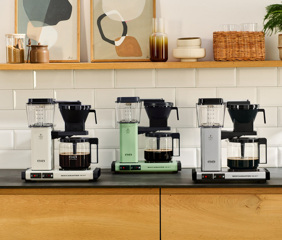 Drei Filterkaffeemaschinen Moccamaster KBG Select, eine weiße, eine mintgrüne und eine silberne, stehen auf einer Küchenarbeitsplatte, jede mit einer Glaskanne Kaffee. Darüber befindet sich ein Holzregal mit Dekorationsartikeln.