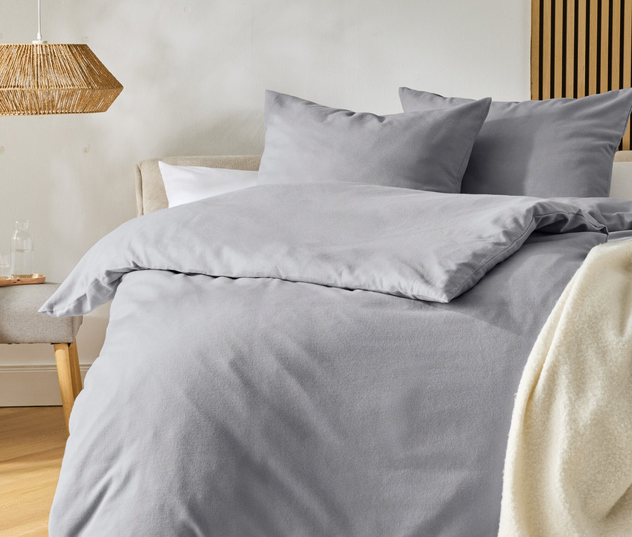 Ein Bett mit hellgrauer Bettwäsche und zwei Kissen, dazu eine cremefarbene Decke mit Fransen. Links steht ein Hocker mit einer Glaskaraffe, darüber hängt eine geflochtene Lampe. Der Boden ist aus Holz, ein heller Teppich liegt davor.