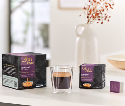 Auf einem Tisch stehen Qbo Espresso ORO TOLIMA Kapselpackungen (27 und 8 Kapseln) neben einem Glas Espresso und einer einzelnen Kapsel.