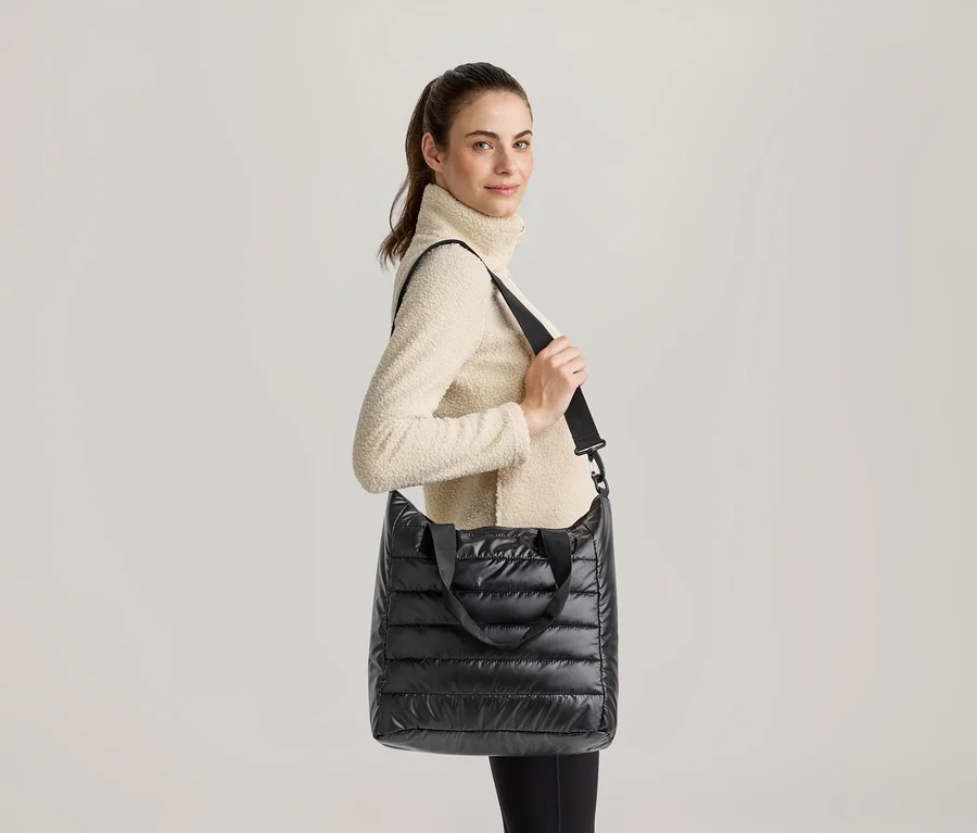 Eine Frau mit Pferdeschwanz, die einen cremefarbenen Fleece-Rollkragenpullover und schwarze Leggings trägt, hält eine schwarze gesteppte Tragetasche mit Schultergurt und blickt lächelnd in die Kamera.