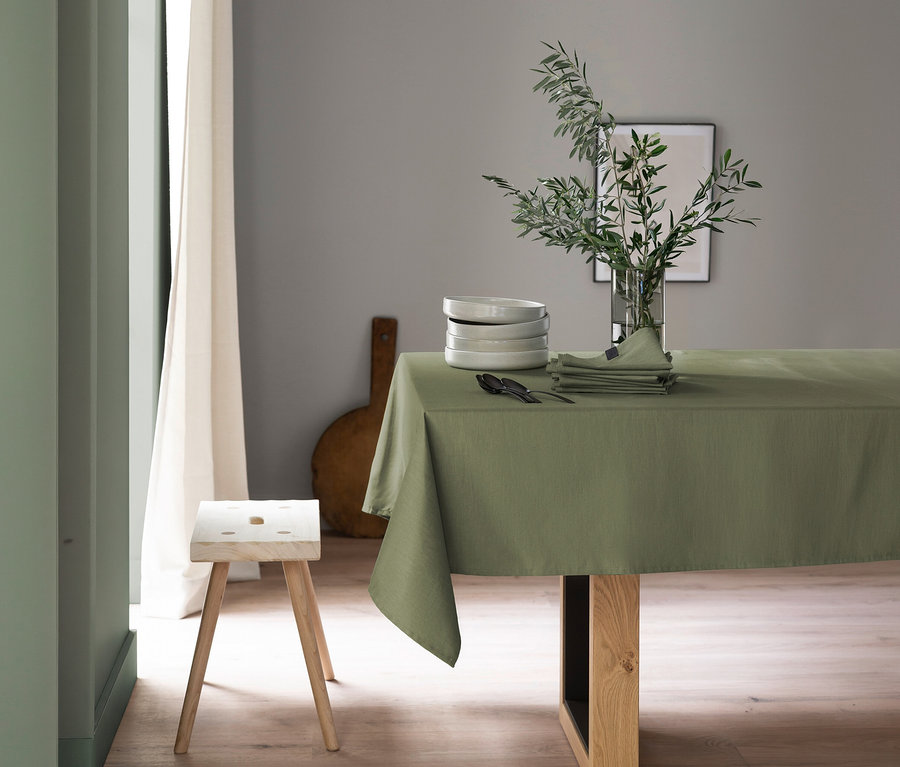 Ein Esstisch ist mit einer khakifarbenen SCHÖNER WOHNEN-Kollektion Tischdecke bedeckt und mit einem Stapel weißer Teller, dunklem Besteck und gefalteten grünen Servietten gedeckt. Eine klare Vase mit Olivenzweigen steht daneben. Im Vordergrund links steht ein heller Holzschemel, und ein Fenster mit Vorhang ist sichtbar.