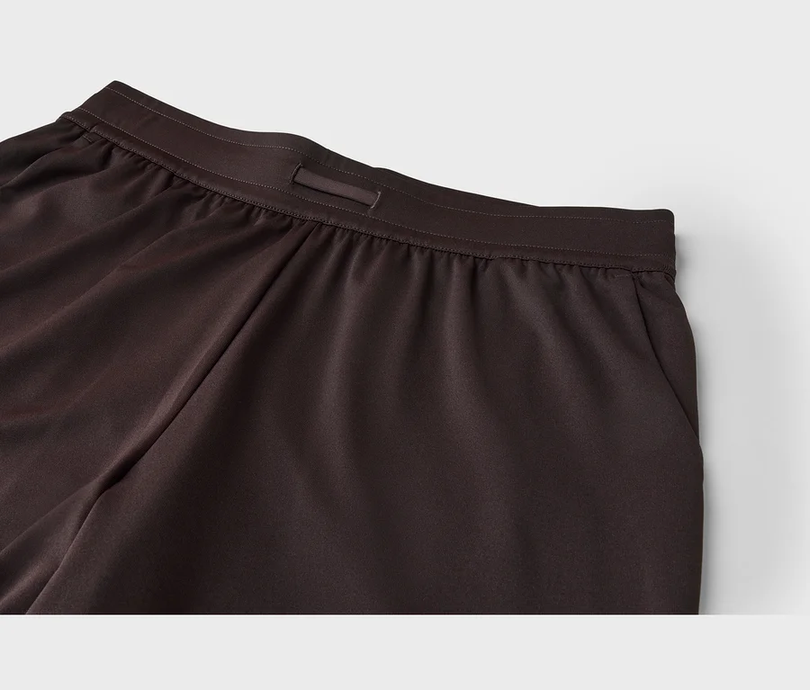 Nahaufnahme einer dunkelbraunen Hose, die den elastischen Bund mit einer kleinen Schlaufe in der vorderen Mitte zeigt. Der glatte Stoff weist leichte Raffungen unterhalb des Bundes auf, eine Taschenöffnung ist rechts sichtbar.