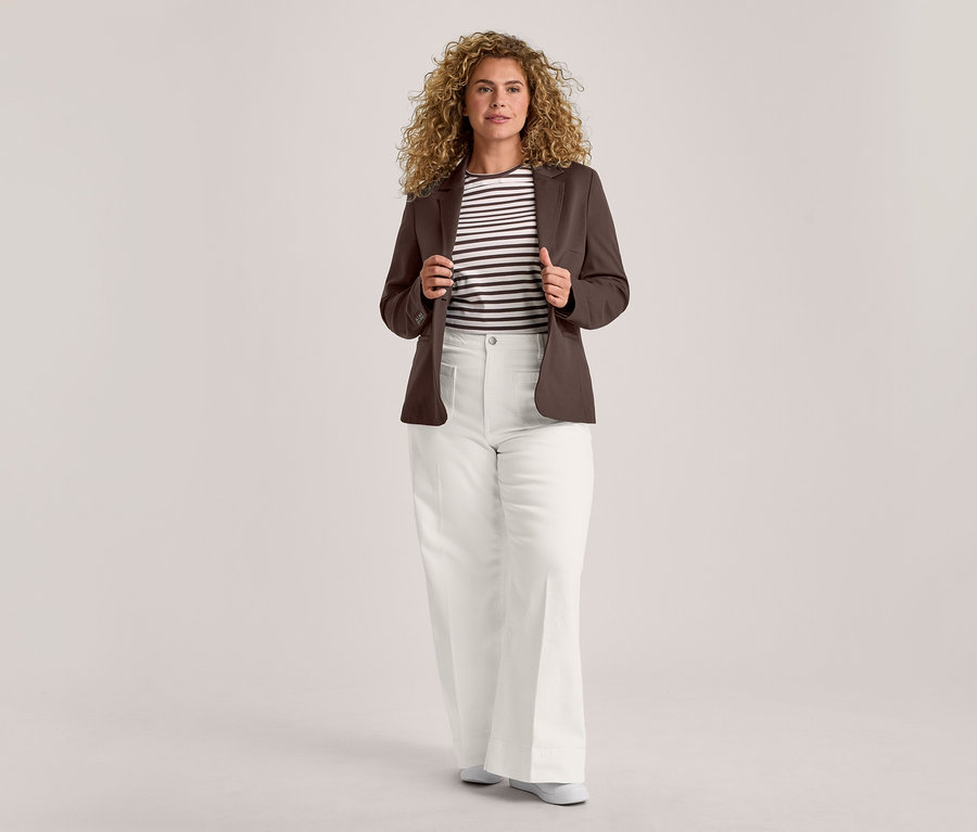 Eine Frau mit lockigem Haar steht frontal, hält die Revers ihres offenen braunen Blazers. Sie trägt ein schwarz-weiß gestreiftes T-Shirt, eine weiße weite Hose und helle flache Schuhe.
