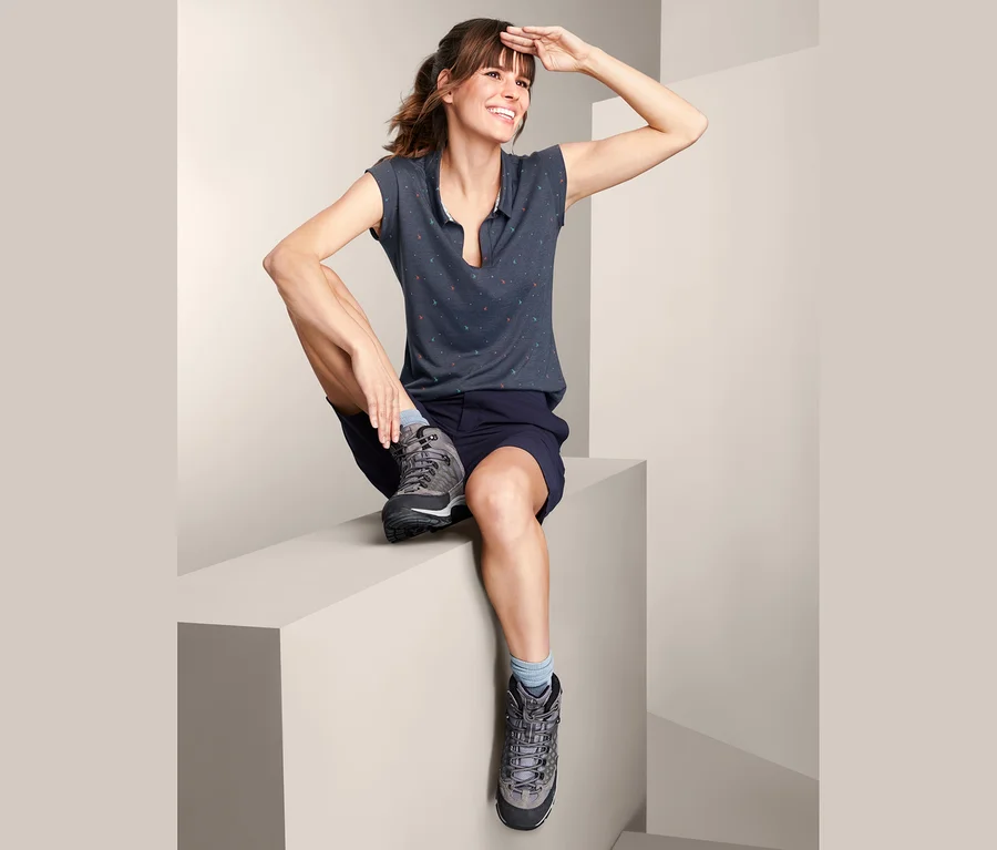 Eine Frau sitzt auf einer Kante und schaut in die Ferne, sie trägt ein Funktionstop, Shorts und Wanderschuhe.