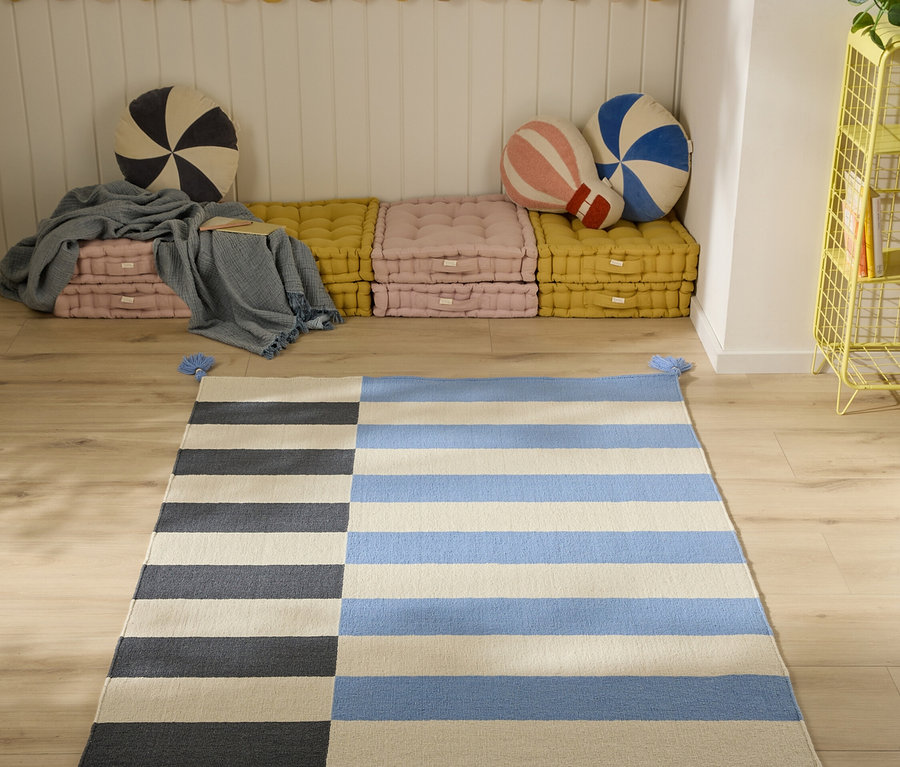 Ein blau, beige und schwarz gestreifter Teppich mit Quasten auf einem Holzfußboden. Dahinter gestapelte rosa und gelbe Bodenkissen mit Zierkissen an einer weißen Paneelwand.