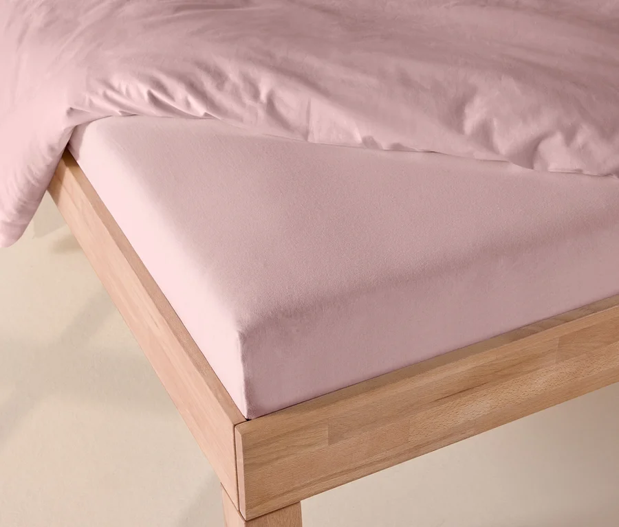 Rosafarbenes Jersey-Spannbettlaken auf einem Bett mit hellem Holzrahmen, teilweise von einer rosafarbenen Bettdecke bedeckt.