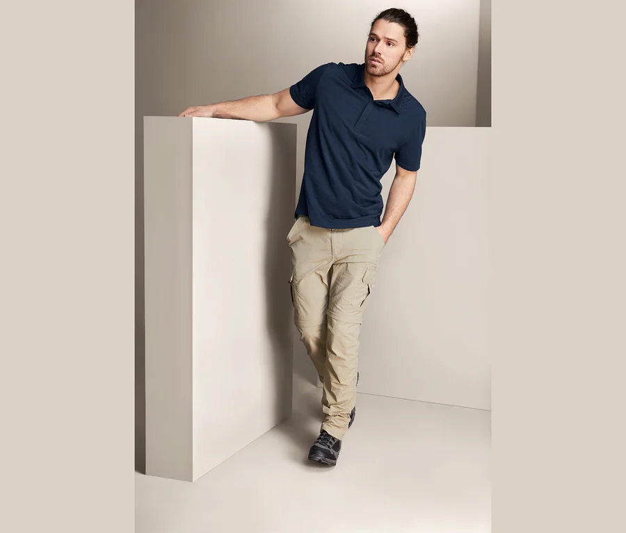 Mann lehnt an einer weißen Wand und trägt ein dunkelblaues Funktions-Poloshirt mit Merinowolle und beige Hosen.