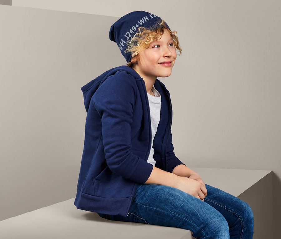 Ein Junge sitzt und trägt eine Wendemütze und eine blaue Kapuzenjacke.